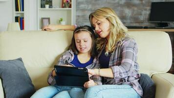 schön jung Mutter mit Tablette zum ihr Tochter Bildung Sitzung auf das Couch im Leben Zimmer. video