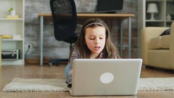 poco ragazza con bretelle dire bugie su il pavimento nel vivente camera utilizzando sua il computer portatile. contento bambino. video