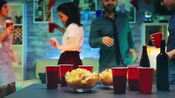 joven hombre tomando papas fritas desde el mesa mientras su amigos son bailando a el fiesta en un habitación con neón luces y disco pelota video