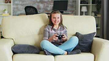 alegre pequeno menina jogando vídeo jogos usando sem fio controlador sentado em a sofá. video