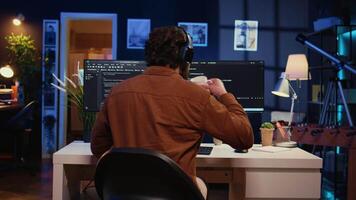 Man doing IT support job from apartment office, drinking cup of coffee and listening music while coding. Developer with headphones on enjoying hot tea and writing code on computer, camera B close up video