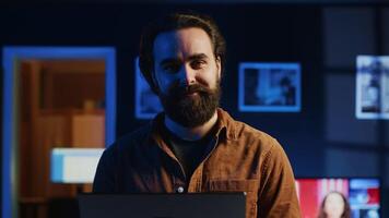 Portrait of happy man doing IT support job from apartment, standing in personal office with laptop in hand, coding. Joyful developer writing lines of code on notebook, camera A close up shot video