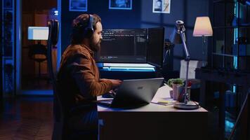 Technician focused on typing complex code in neon lit home office, developing software application. It specialist removing headphones to better concentrate on difficult task, camera B video