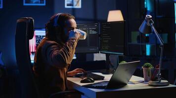 Programmer does multitasking to finish developing PC code, drinks coffee, listens music. IT admin wearing headphones, enjoying hot beverage, solving database errors on computer and laptop, camera A video