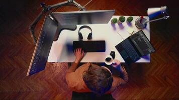 Top down view of engineer doing multitasking to finish developing PC code, drinking coffee, listening music. IT admin wearing headphones, solving database errors on computer and laptop, aerial shot video