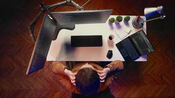 Aerial shot of computer scientist listening music while writing lines of code during remote job shift in home office. IT professional enjoying audio podcast, coding on desktop PC, top down view video