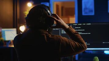 Engineer wearing headphones while using coding to fortify security, remotely working for IT company. Software expert listening music and upgrading binary code scripts on computer, camera A video