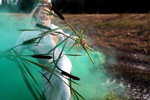 Cheerful young  woman with reeds dances in colored smoke in a field photo