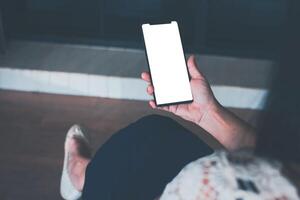 mujer participación un teléfono inteligente con blanco pantalla Bosquejo, sentado en un silla en el vivo habitación a hogar. Clásico tono foto