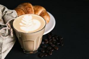 Latte art coffee in a white cup, croissants, roasted bean coffee on black background,  copy space your text photo