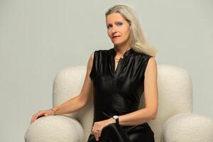 Fashionable elderly woman in stylish black clothes posing on a chair. Studio portrait of an adult blonde. photo