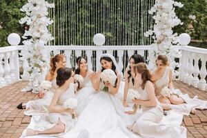 A brunette bride and her bridesmaids in matching cream dresses sit with the bride and rejoice with flowers in their hands near the ceremonial arch. Wedding in nature. photo
