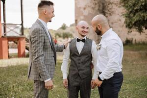 Cheerful, young, energetic witnesses of the groom next to the groom. Friends congratulate the groom. The groom in a vest and his friends in white shirts pose near the wedding arch. Wedding in nature. photo
