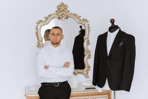 Portrait of a young groom at home before the wedding ceremony. A handsome man is standing near a mirror and looking at the camera. photo