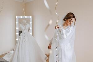 A beautiful bride with a long veil in her room, wearing a robe. Wedding dress on a mannequin. The bride in the morning before the wedding ceremony. photo