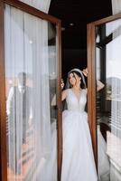 el novia soportes por el ventana en un blanco vestir y mira a el novio. Boda día. maravilloso novia. casamiento. foto