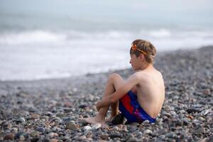 A little boy sits on a stone shore and looks at the sea. photo