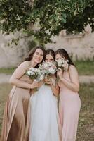 A brunette bride and her bridesmaids in cream dresses stand and cover their faces with bouquets of flowers. Girls in matching dresses. Wedding in nature photo