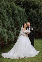 A handsome groom embraces his bride in a lush white dress and smiles in a beautiful outdoor setting. Under the open sky. High quality photo. A newlywed couple poses together on a sunny summer day. photo