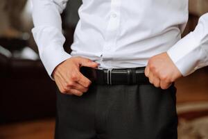 Close-up of a man holding a black leather belt. A man in a white shirt, without a face, holds a black leather belt in his hands. The morning of the groom at the wedding. photo