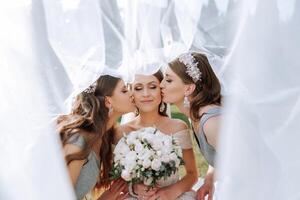 Girlfriends kiss the bride on the cheeks, wrapped in a veil. The bride is holding a bouquet. Girls in identical dresses photo