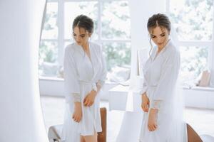 novia en un Boda vestir mirando en el espejo. hermosa joven niña novia en el estudio cerca el espejo en un blanco túnica. foto