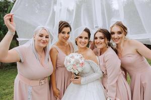 Bridesmaids are wrapped in a veil. The bride is holding a bouquet. Long veil. Wedding in nature. Smiling girls in identical dresses. photo