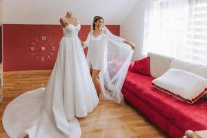 un hermosa novia con un largo velo en su habitación, vistiendo un túnica. Boda vestir en un maniquí. el novia en el Mañana antes de el Boda ceremonia. foto