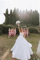 A brunette bride and her bridesmaids in pink dresses are walking in the garden. A long train on the dress. Wedding in nature photo