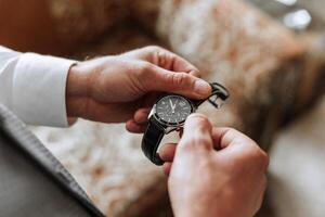 A man in a business suit holds a wristwatch in his hands, a mug of coffee is on the table in his room photo