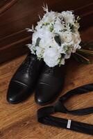 On the floor are men's dark leather shoes and a black belt, a man's suit on a mannequin, a wedding bouquet of flowers, wedding rings, men's perfume. Photo, top view. photo