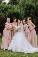A brunette bride and her bridesmaids in identical pink dresses are standing against the background of nature. Girls in identical dresses are making out at a wedding. Wedding in nature. photo