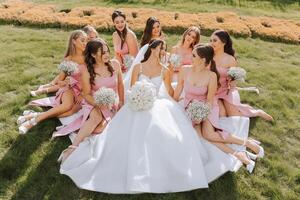 A brunette bride and her bridesmaids in identical dresses sit with the bride and rejoice with flowers in their hands on the green grass. Wedding in nature. photo