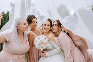 Bridesmaids are wrapped in a veil. The bride is holding a bouquet. Long veil. Wedding in nature. Smiling girls in identical dresses. photo
