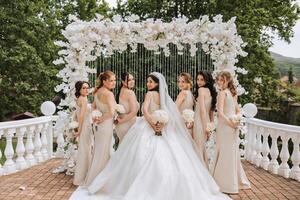 The bride and her bridesmaids turned their backs to the camera against the background of green trees. A long train on the dress. The girls are wearing identical dresses at the wedding. photo