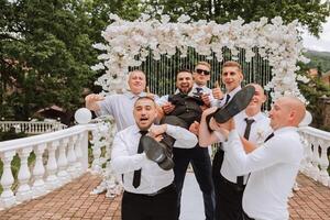 Cheerful, young, energetic witnesses of the bridegroom hold the bridegroom in their arms. Funny photo. The groom in a vest and his friends in white shirts pose near the wedding arch. Wedding in nature photo