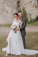 un hermoso novio abraza su novia en un lozano blanco vestir y sonrisas en un hermosa al aire libre ajuste. debajo el abierto cielo. alto calidad foto. un recién casado Pareja poses juntos en un soleado verano día. foto