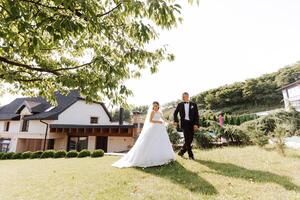 A handsome groom and an elegant bride in a lush white dress are walking in a summer park. Happy bride and groom getting ready for their best day. photo