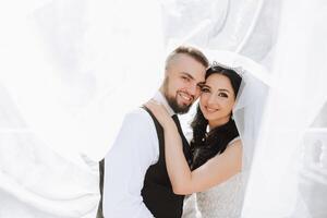 un hermoso novio abraza su novia en un lozano blanco vestir y sonrisas en un hermosa al aire libre ajuste. debajo el abierto cielo. alto calidad foto. un recién casado Pareja poses juntos en un soleado verano día. foto