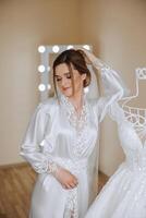 A beautiful bride with a long veil in her room, wearing a robe. Wedding dress on a mannequin. The bride in the morning before the wedding ceremony. photo