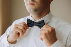 A bearded man in a white shirt adjusts his bow tie. Groom's morning. Close-up detail, men's tie for a wedding or an important meeting. photo