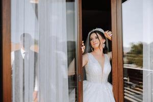 el novia soportes por el ventana en un blanco vestir y mira a el novio. Boda día. maravilloso novia. casamiento. foto