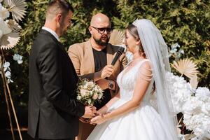 Boda ceremonia en naturaleza. el novia y novio cerca el flor arco. Maestro de ceremonias en oscuro lentes a un Boda durante un actuación en contra el antecedentes de el novia y novio. foto