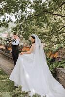 A handsome groom and an elegant bride in a lush white dress are walking in a summer park. Happy bride and groom getting ready for their best day. photo