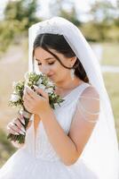un encantador novia en un increíblemente hermosa vestir disfruta un ramo de flores y olfatea él. verano boda. un lujoso novia. foto