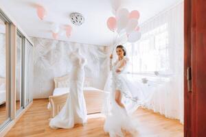 A beautiful bride with a long veil in her room, wearing a robe. Wedding dress on a mannequin. The bride in the morning before the wedding ceremony. photo