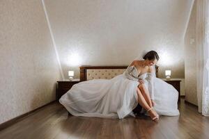 joven novia en hermosa Boda vestir poniendo en Zapatos adentro. novia vestidos Zapatos antes de el Boda ceremonia. detalle de novia poniendo en alto bien portado sandalia Boda zapatos. Boda novia zapatos. foto