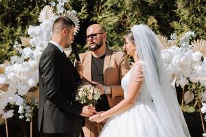 Boda ceremonia en naturaleza. el novia y novio cerca el flor arco. Maestro de ceremonias en oscuro lentes a un Boda durante un actuación en contra el antecedentes de el novia y novio. foto