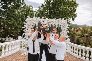 Cheerful, young, energetic witnesses of the bridegroom hold the bridegroom in their arms. Funny photo. The groom in a vest and his friends in white shirts pose near the wedding arch. Wedding in nature photo