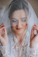 Beautiful young bride holding veil in white wedding dress, portrait of brunette bride in hotel room, morning before wedding. photo